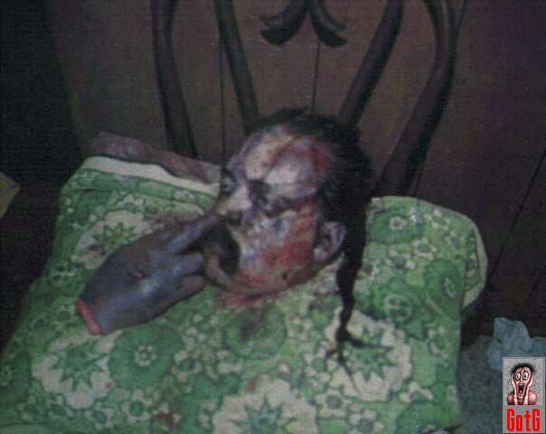 Severed head with severed hand on chair with finger in nose.